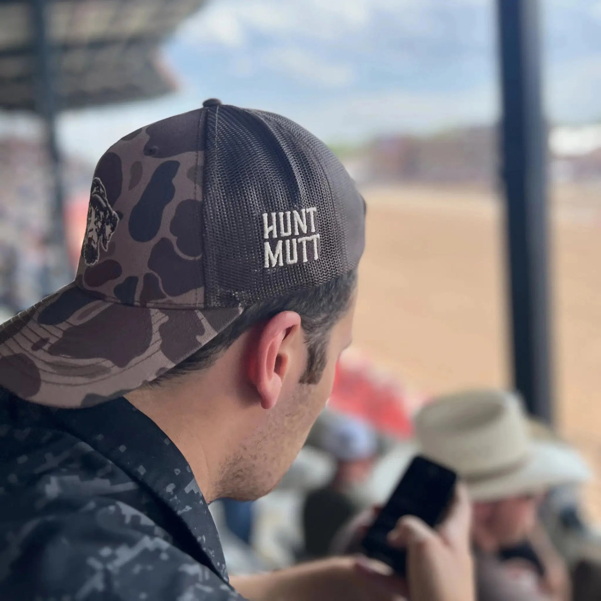 Person wearing a cap with 'Hunt Mutt' logo, looking at a phone at a rodeo